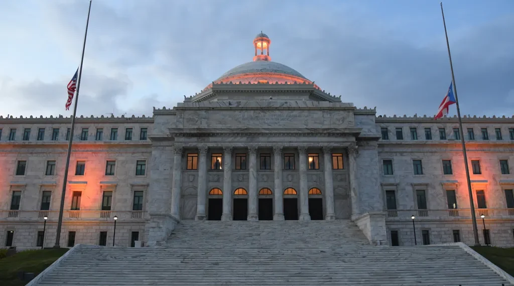 The Capitol of Puerto Rico overlooks San Juan Bay at dusk with the United States and Puerto Rico flags flying at half-staff.