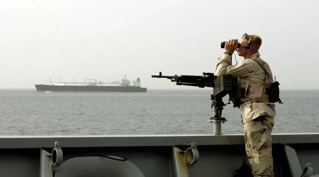A U.S. Navy sailor stands watch aboard a military vessel while transiting the Strait of Hormuz, monitoring nearby maritime traffic.