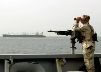 A U.S. Navy sailor stands watch aboard a military vessel while transiting the Strait of Hormuz, monitoring nearby maritime traffic.