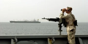 A U.S. Navy sailor stands watch aboard a military vessel while transiting the Strait of Hormuz, monitoring nearby maritime traffic.