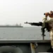 A U.S. Navy sailor stands watch aboard a military vessel while transiting the Strait of Hormuz, monitoring nearby maritime traffic.