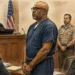 Courtroom illustration showing a handcuffed defendant standing before a judge, with attorneys, a law enforcement officer and observers present.