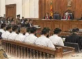Courtroom hearing shows judges seated at the bench, defendants facing forward, lawyers and police officers present during proceedings.