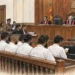 Courtroom hearing shows judges seated at the bench, defendants facing forward, lawyers and police officers present during proceedings.