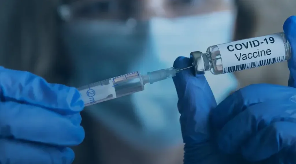 A gloved person wearing a surgical mask handles a COVID-19 vaccine vial and syringe during vaccination preparation.