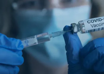 A gloved person wearing a surgical mask handles a COVID-19 vaccine vial and syringe during vaccination preparation.