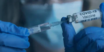 A gloved person wearing a surgical mask handles a COVID-19 vaccine vial and syringe during vaccination preparation.