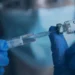 A gloved person wearing a surgical mask handles a COVID-19 vaccine vial and syringe during vaccination preparation.