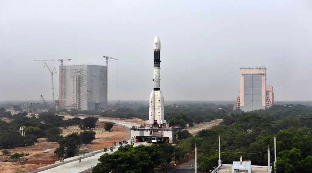 GSLV-F08 rocket stands on a mobile launch pedestal with the Vehicle Assembly Building visible behind it at an ISRO launch complex.