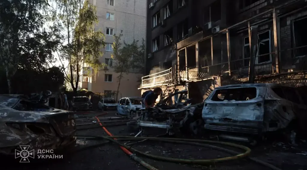 Damaged apartment building and burned vehicles at a Kyiv attack site as a person works among debris and emergency hoses.