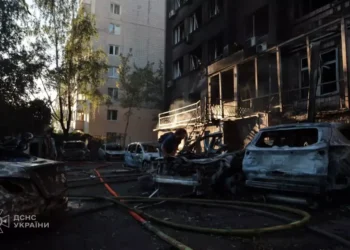 Damaged apartment building and burned vehicles at a Kyiv attack site as a person works among debris and emergency hoses.
