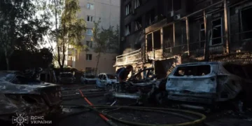 Damaged apartment building and burned vehicles at a Kyiv attack site as a person works among debris and emergency hoses.