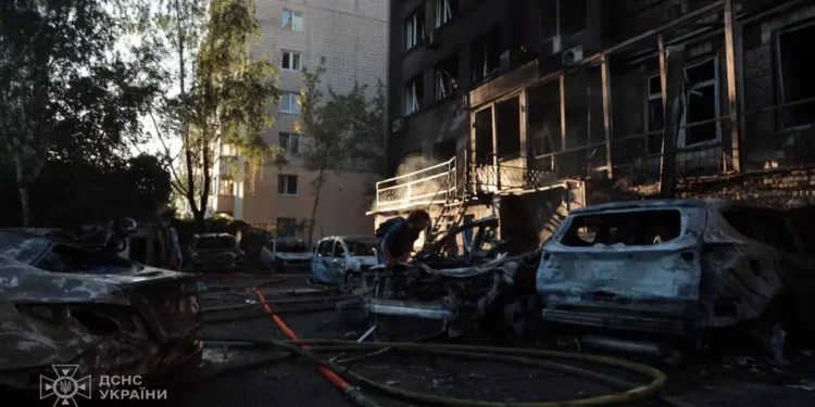 Damaged apartment building and burned vehicles at a Kyiv attack site as a person works among debris and emergency hoses.