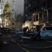 Damaged apartment building and burned vehicles at a Kyiv attack site as a person works among debris and emergency hoses.
