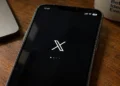 Mobile phone displays the X app loading screen on a wooden desk beside a laptop and coffee mug.