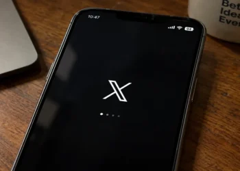 Mobile phone displays the X app loading screen on a wooden desk beside a laptop and coffee mug.