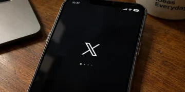 Mobile phone displays the X app loading screen on a wooden desk beside a laptop and coffee mug.