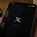 Mobile phone displays the X app loading screen on a wooden desk beside a laptop and coffee mug.