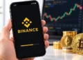 A smartphone displays the Binance logo and mobile app loading screen, with Bitcoin and Ethereum coins, stacks of cryptocurrency tokens, and a digital trading chart in the background.