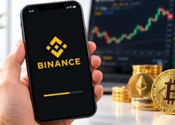 A smartphone displays the Binance logo and mobile app loading screen, with Bitcoin and Ethereum coins, stacks of cryptocurrency tokens, and a digital trading chart in the background.