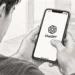 A person holds a smartphone displaying the ChatGPT logo in a pencil-sketch illustration, with a notebook, pen and plant on a desk.