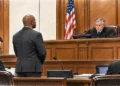 A courtroom illustration shows a defendant standing before a judge at an elevated wooden bench, with a uniformed sheriff and attorneys present during a U.S. court hearing.