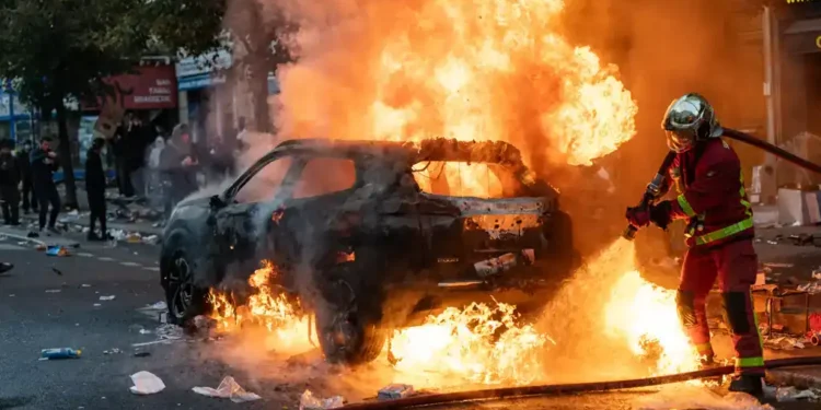 Firefighters extinguish a burning car during protests and riots in Paris, France, on October 17, 2026, amid smoke and unrest.